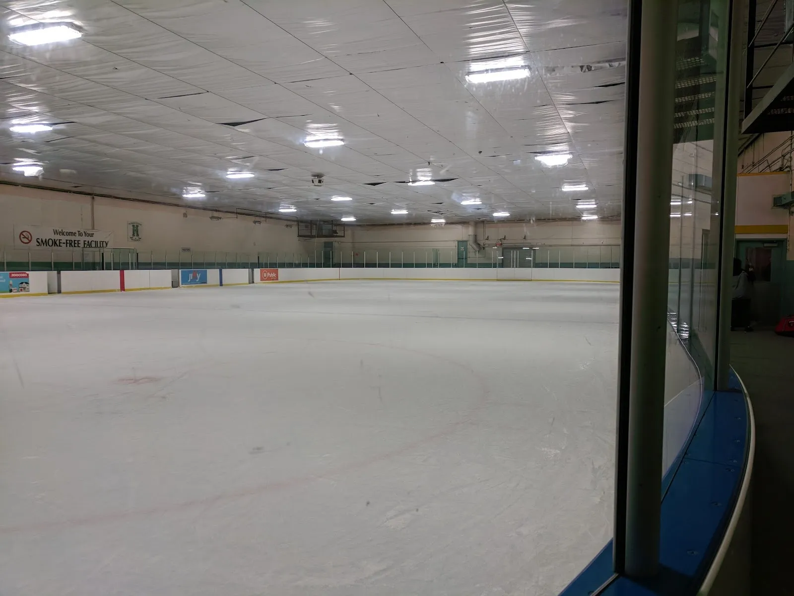 Una pista amplia de aspecto vacío vista junto al vidrio, con la línea azul y un techo gris claro con largas luminarias sobre las vallas del fondo. — John Booth Memorial Arena