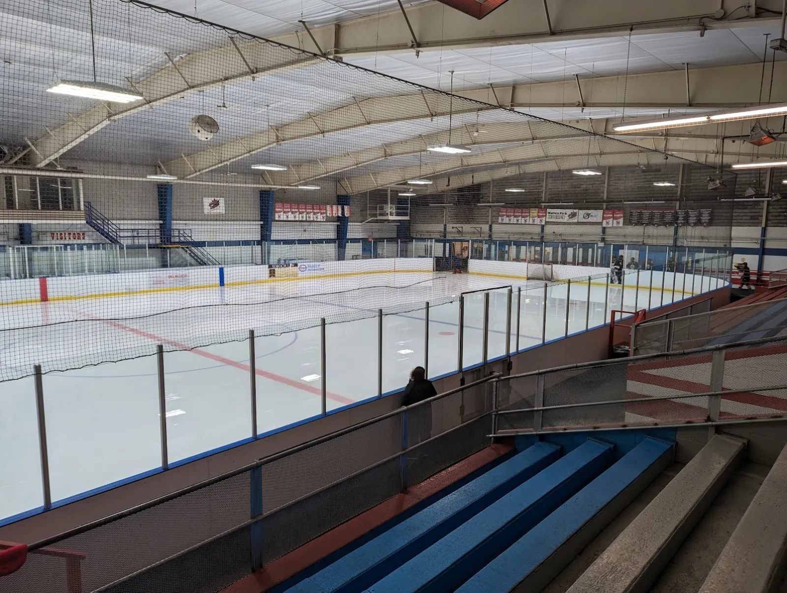 Full ice sheet at Lambton Arena seen from an elevated corner of the stands, with the blue-seat bleachers, a "VISITORS" sign, and team banners along the boards; one bystander stands at the glass. — Lambton Arena