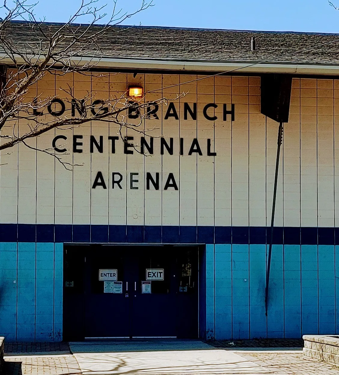 The front of the Long Branch Centennial Arena on Arcadian Circle in Toronto, with its lettered name above the blue entrance doors — Long Branch Arena