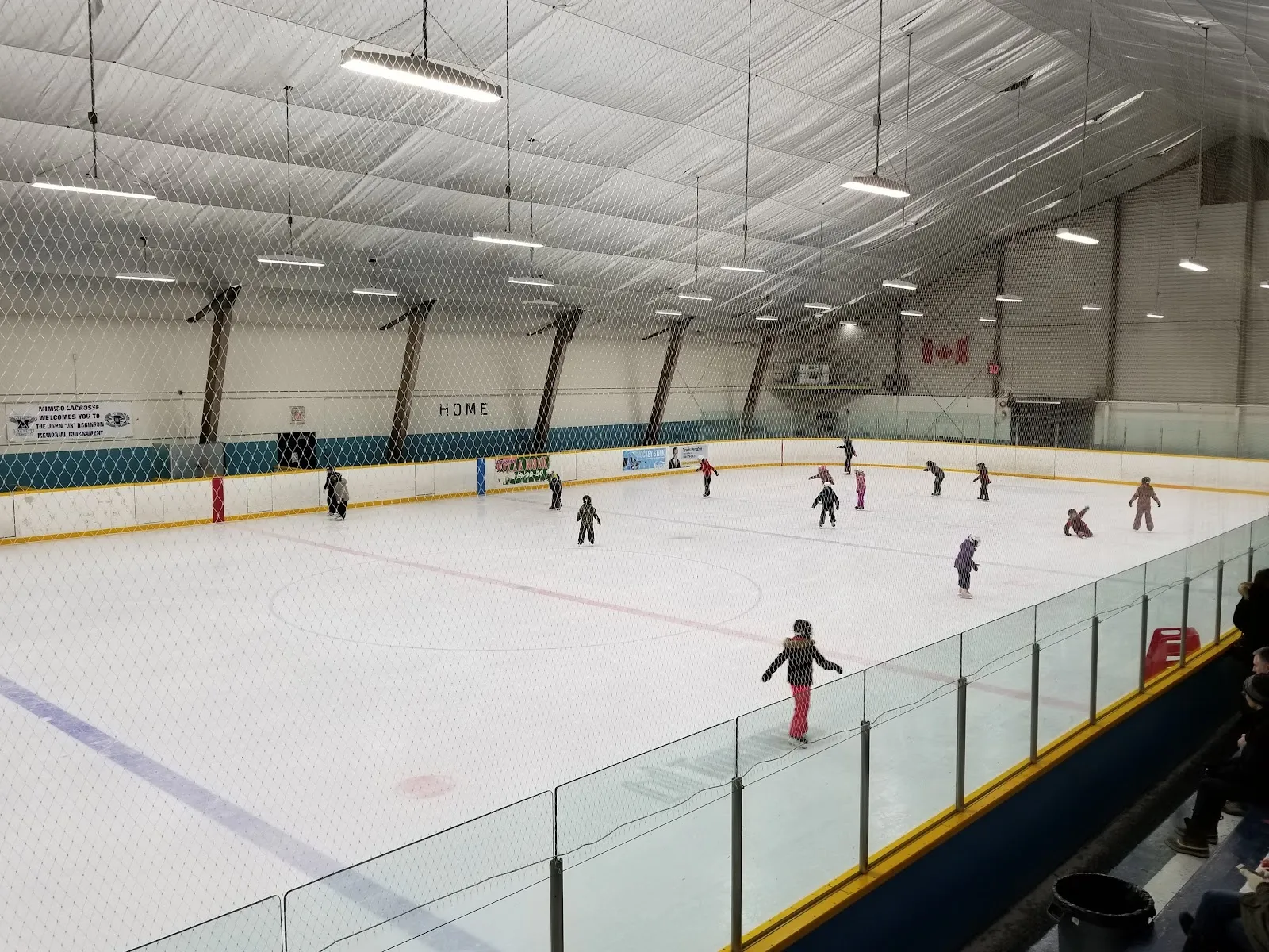 Skaters spread across a wide sheet in a public session seen from the stands past a clear rail, white ceiling panels and a long arched roof above. — Long Branch Arena