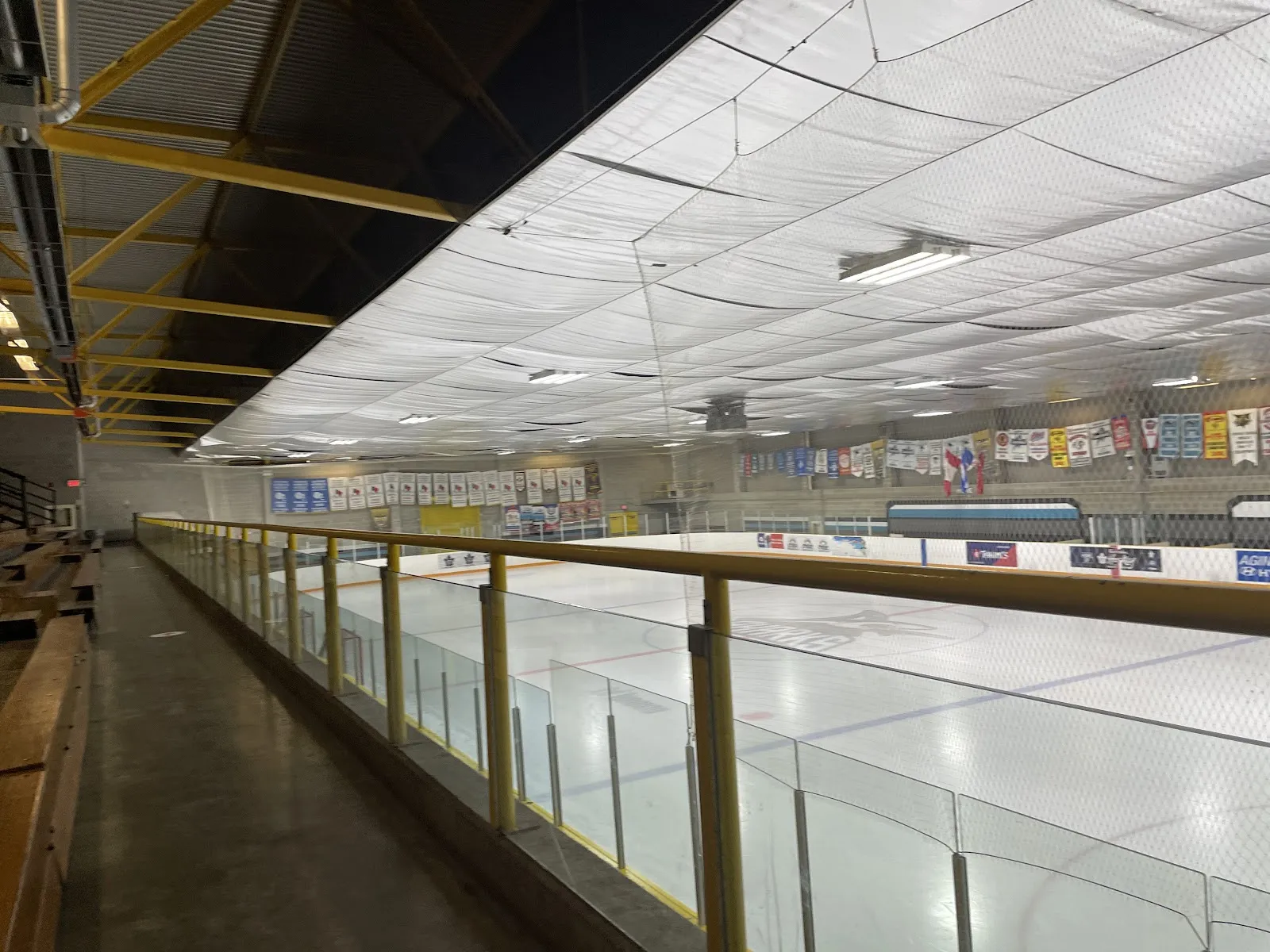 El hielo vacío del Malvern Community Recreation Centre visto a lo largo de la baranda desde el pasillo superior, bajo el revestimiento blanco del techo y las mantas de los clubes — Malvern Recreation Centre