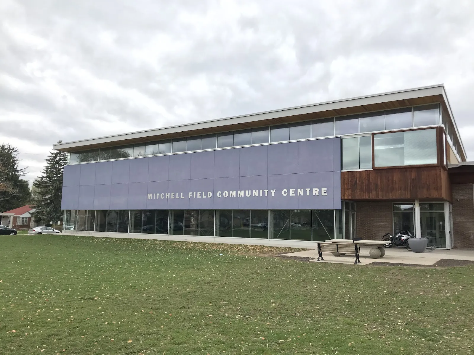 Front entrance corner of Mitchell Field Community Centre, brick and dark-wood cladding with a large glazed entry bay, parked motorcycle and bench out front, no people. — Mitchell Field Community Centre