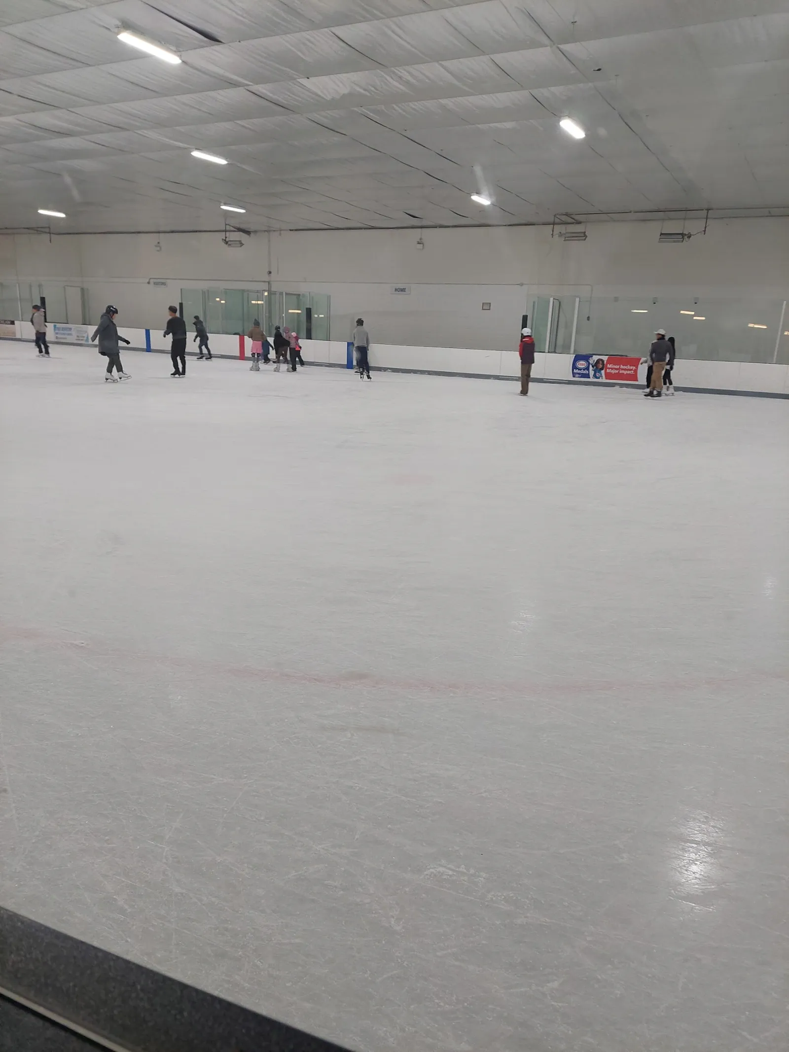 Patinaje público visto a nivel del hielo, con algunos patinadores cerca de las vallas del fondo bajo un techo blanco con largas lámparas lineales. — Oriole Community Recreation Centre
