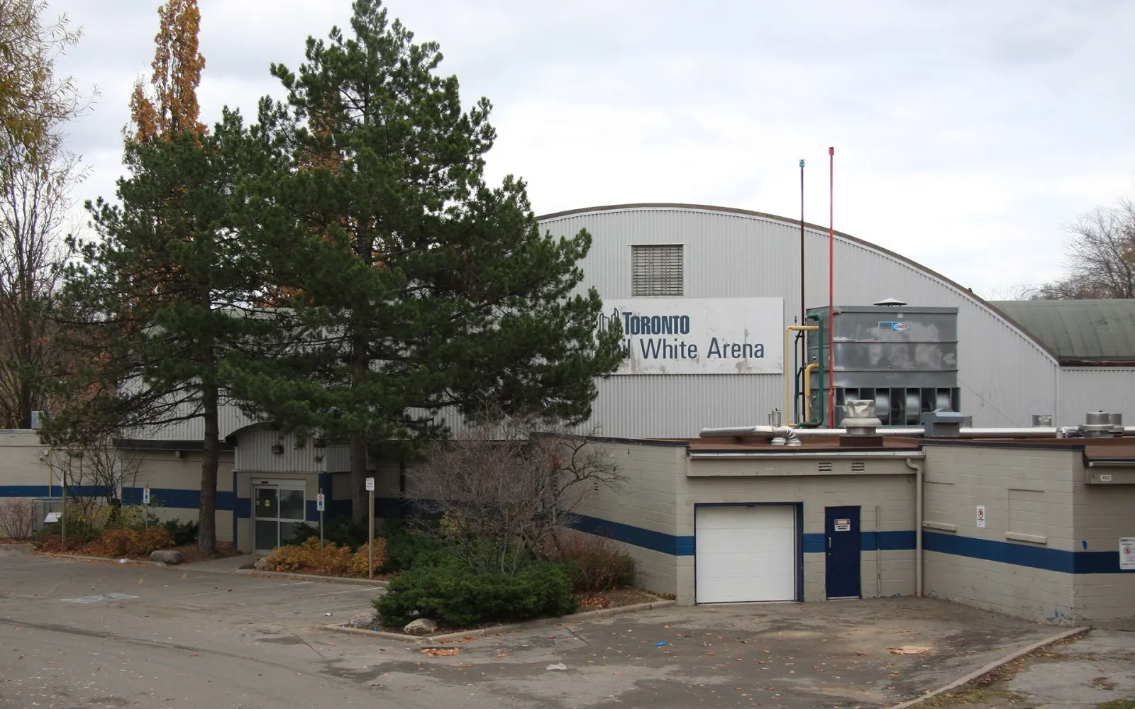 Phil White Arena, a barrel-roofed building with a blue stripe along its base, seen through trees from the parking area. — Phil White Arena