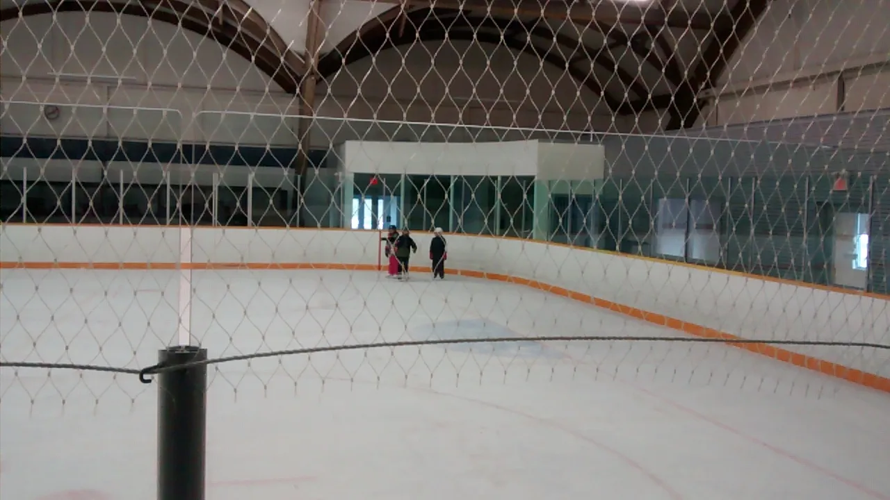 The ice at Phil White Arena behind the safety netting, under the arched timber roof, with orange boards and three small figures far across the rink — Phil White Arena