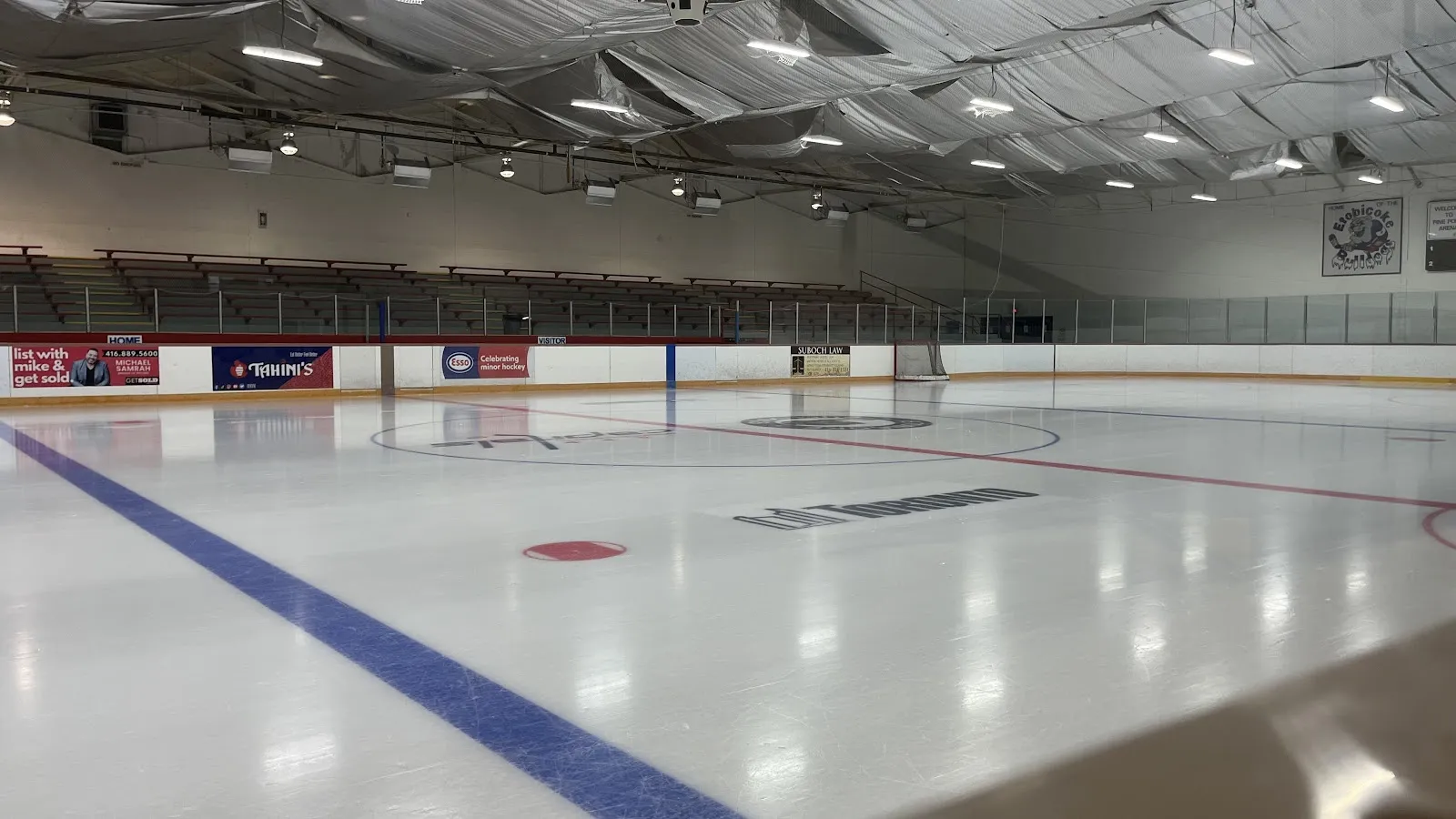 Empty ice sheet from ice level, boards lined with local-business ads, an "Etobicoke" crest visible on the far wall. — Pine Point Arena