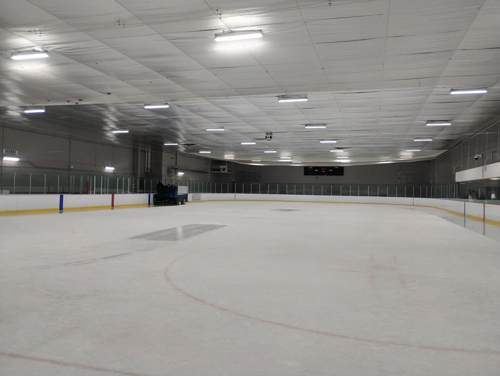 Pista vacía recién pulida desde el nivel del hielo mirando hacia el marcador y el área de espectadores, con una pulidora de hielo estacionada junto a las vallas del fondo. — Pleasantview Community Centre