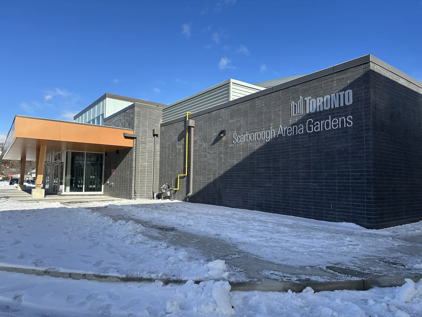 Esquina del edificio de Scarborough Arena Gardens en un día despejado de invierno, con revestimiento de ladrillo oscuro, el logotipo de 'Toronto' en relieve y las letras de 'Scarborough Arena Gardens', la entrada con marquesina a la izquierda, nieve en el suelo y sin personas. — Scarborough Arena Gardens