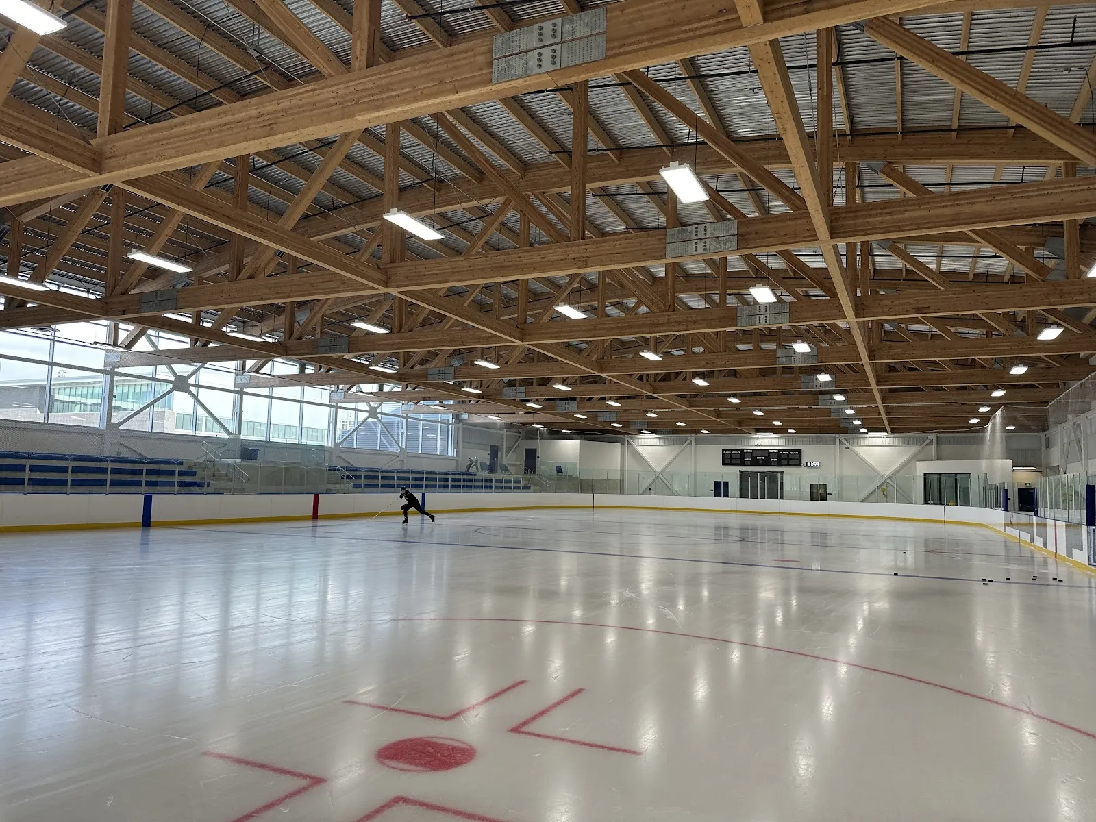 Amplia vista interior de una pista vacía en Scarborough Arena Gardens bajo un techo de armaduras de madera expuestas, con perímetro de vallas y cristal, asientos para espectadores a un lado, el logotipo de 'City of Toronto' visible en el hielo y un patinador a lo lejos. — Scarborough Arena Gardens