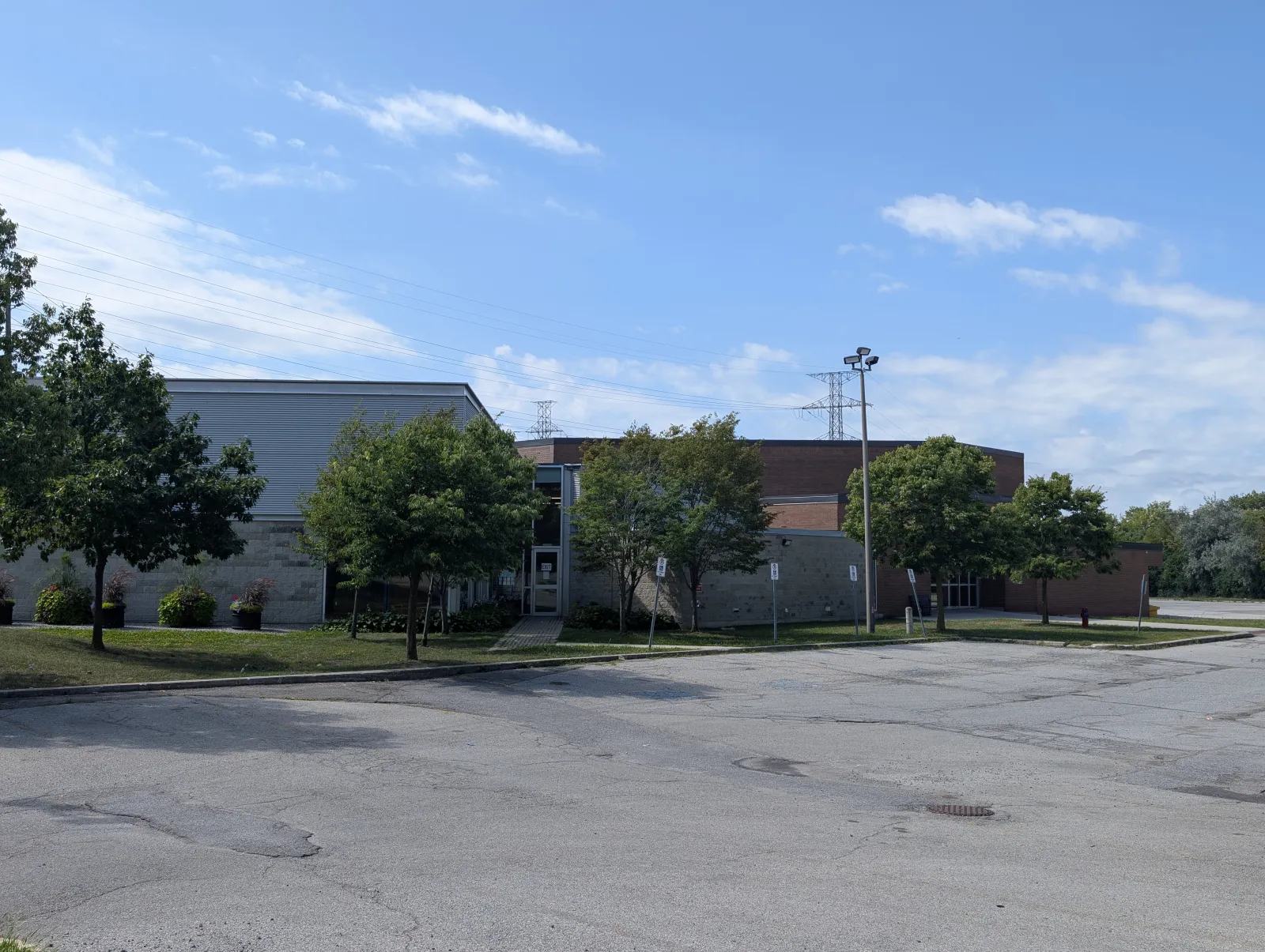 El exterior del centro recreativo desde el estacionamiento: un edificio de piedra y ladrillo con un ala de la arena de techo curvo de metal acanalado, un vestíbulo de entrada de vidrio y árboles y postes de luz a lo largo del andador. — Victoria Village Recreation Centre and Arena