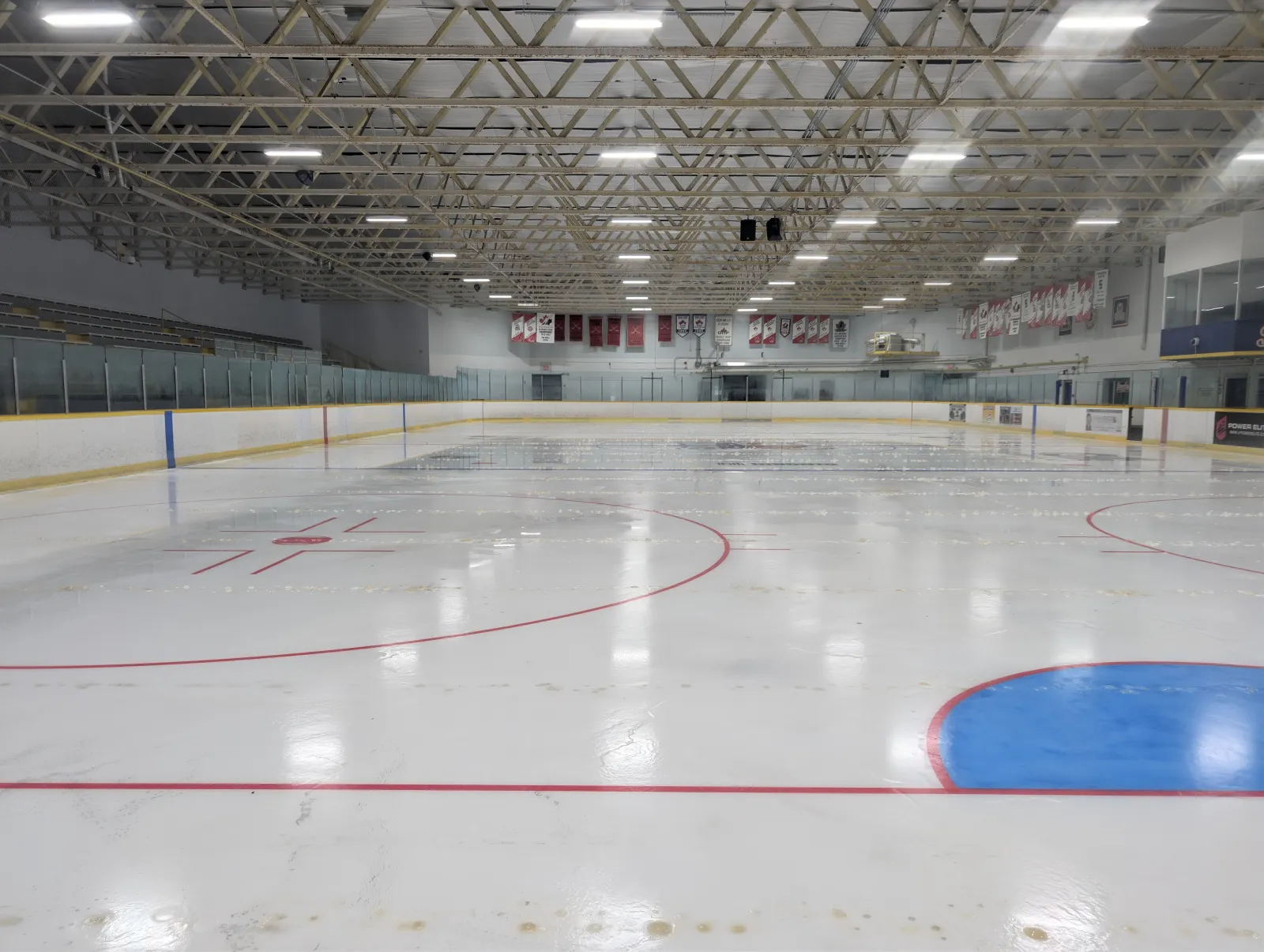 Pista vacía recién pulida dentro de la arena, vista a nivel del hielo con las vallas, las gradas y mantas de campeonato a lo largo de la pared del fondo. — Victoria Village Recreation Centre and Arena