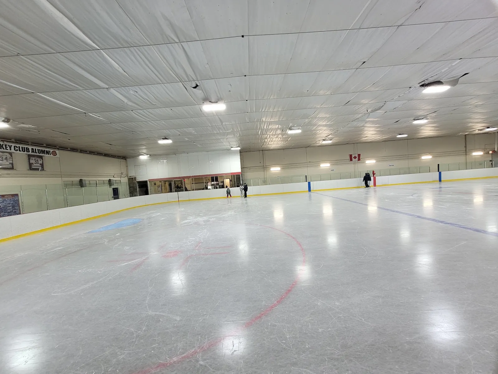 El hielo vacío visto desde la esquina, con vallas de borde amarillo, una bandera de Canadá y un techo blanco bajo con luces redondas. — York Mills Arena