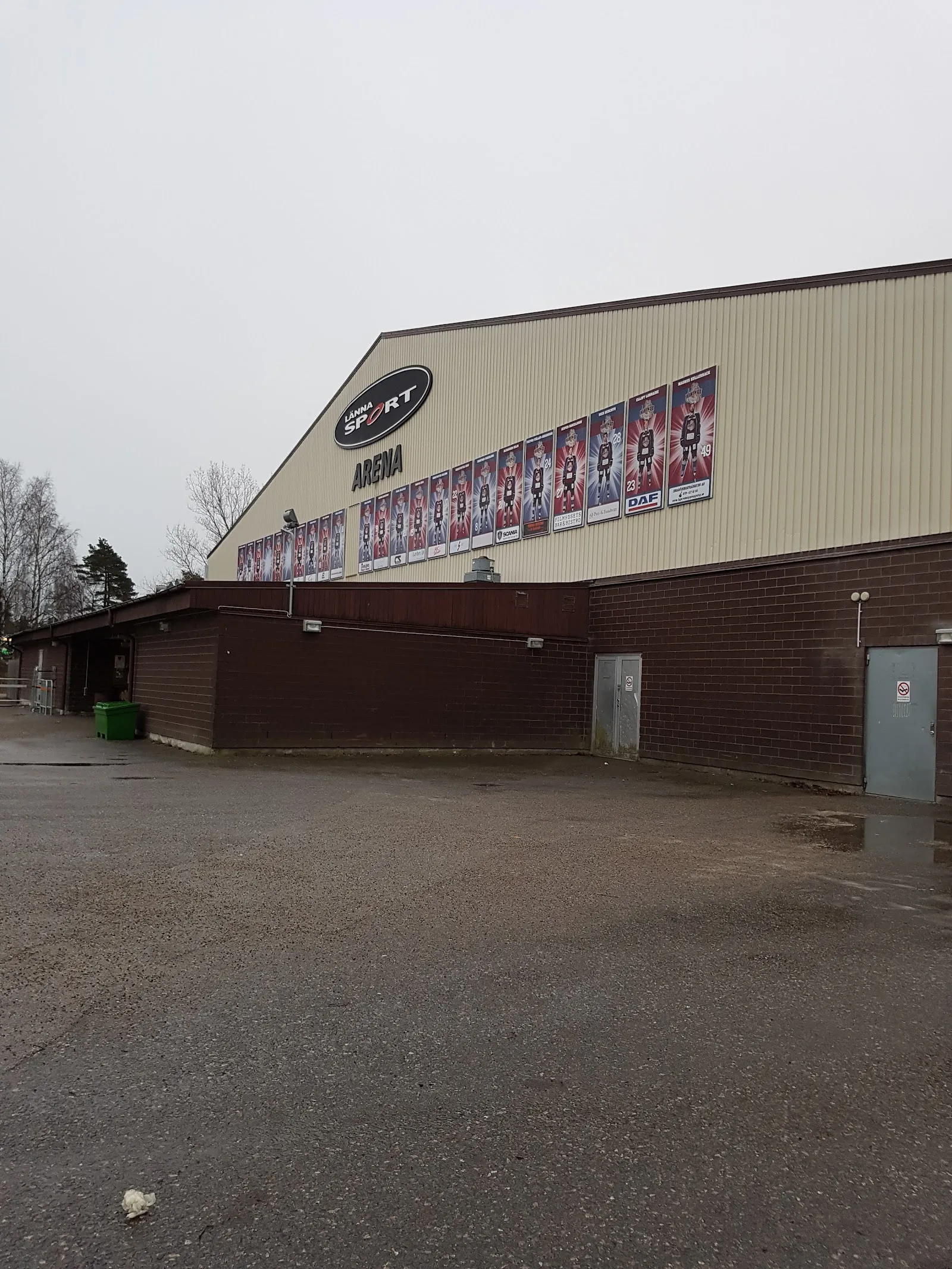 Gavelväggen på Torvalla ishall med raden av banderoller ovanför den bruna sockeln och servicegården – Torvalla ishallar