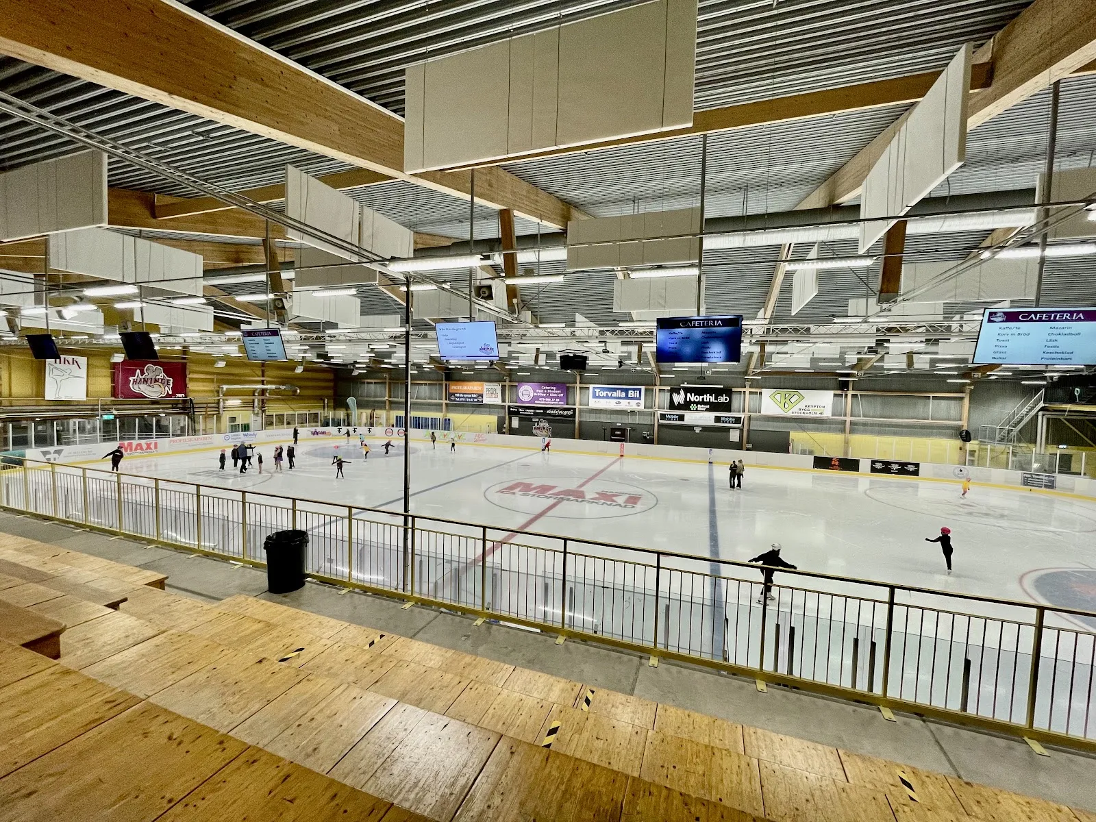 Åkare på isen i Torvalla ishall sedda från träläktaren, med reklamsarg runt rinken – Torvalla ishallar
