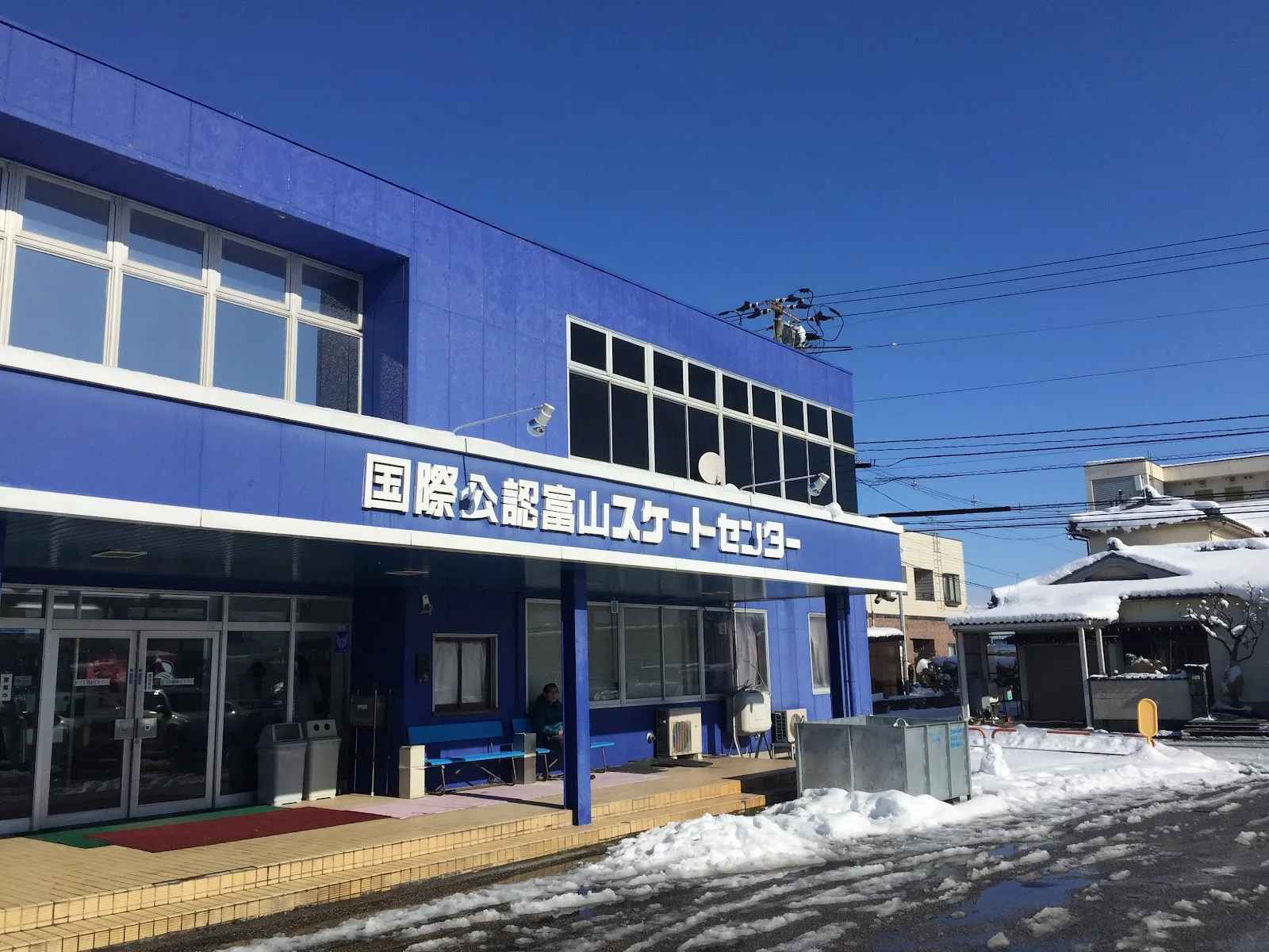 晴れた冬空の下の富山スケートセンターの青い正面。入口の上に看板があり、通路のわきに雪が積まれています。 — Toyama Skate Center