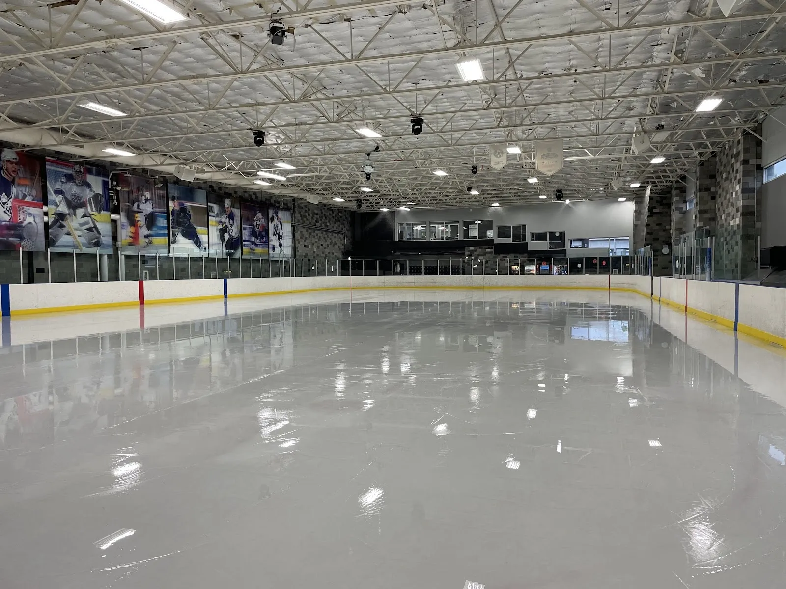 The empty sheet from the end boards, a long wall of team photographs and banners above yellow-edged boards under a steel-trussed roof. — Toyota Sports Performance Center