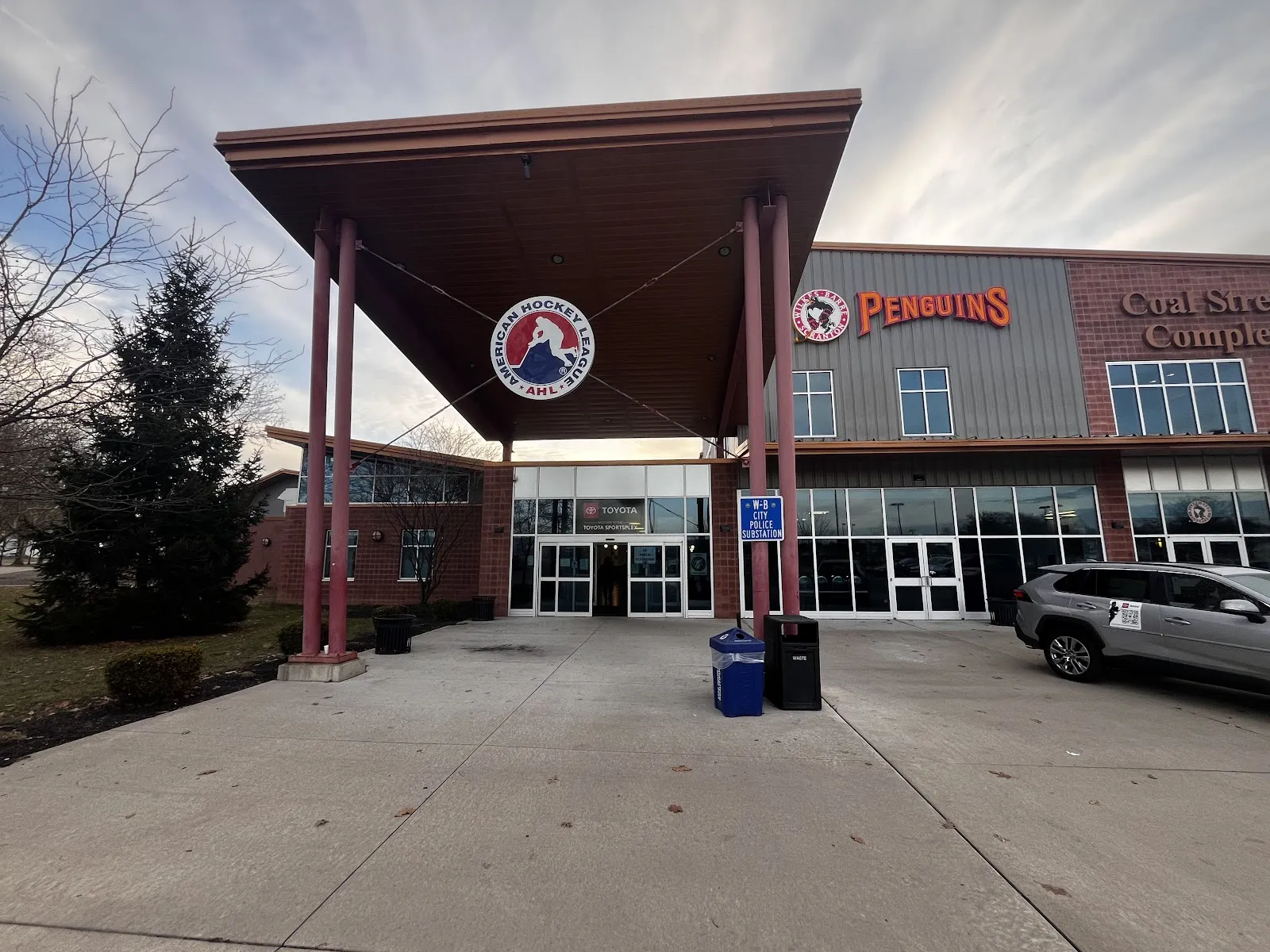 Marquesina de la entrada principal del Toyota SportsPlex (Coal Street Complex) con los logotipos de la AHL y de los Wilkes-Barre/Scranton Penguins montados en el alero puntiagudo, edificio de ladrillo y paneles metálicos, puertas de vidrio en la entrada y un letrero de una subestación de policía. — Toyota SportsPlex