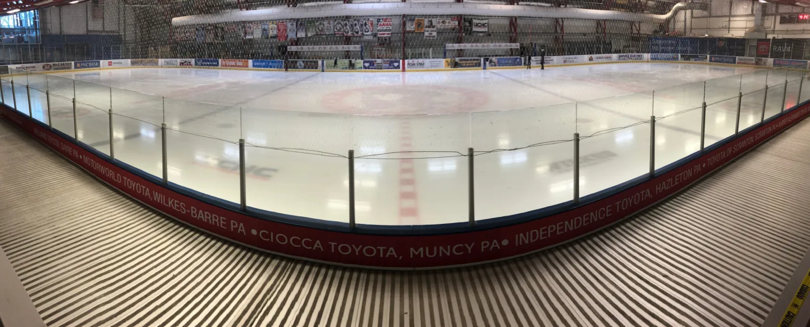 Amplia vista interior de la pista desde junto a la pista, con vallas cubiertas de anuncios de concesionarios Toyota, mantas colgadas de las vigas y dos patinadores a lo lejos en el hielo — Toyota SportsPlex