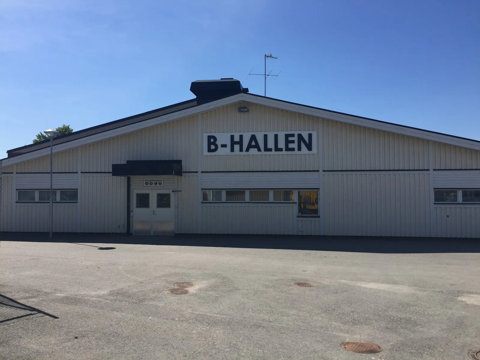 Den ljusa träklädda B-hallen på Trängens idrottsplats med namnet på gaveln, sedd över parkeringen en klar dag – Trängens IP ishall B