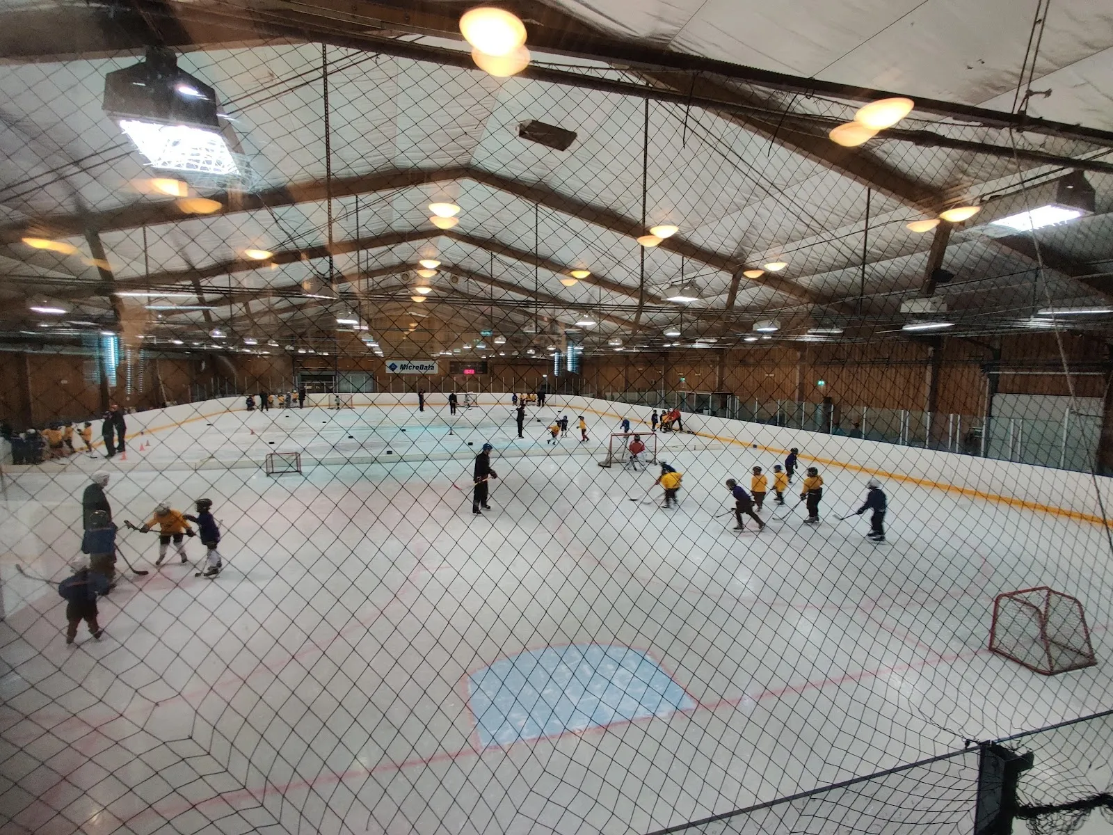 En match pågår på isen i Trängens ishall B i Örebro, sedd genom nätet över hela isytan under trätaket. – Trängens IP ishall B