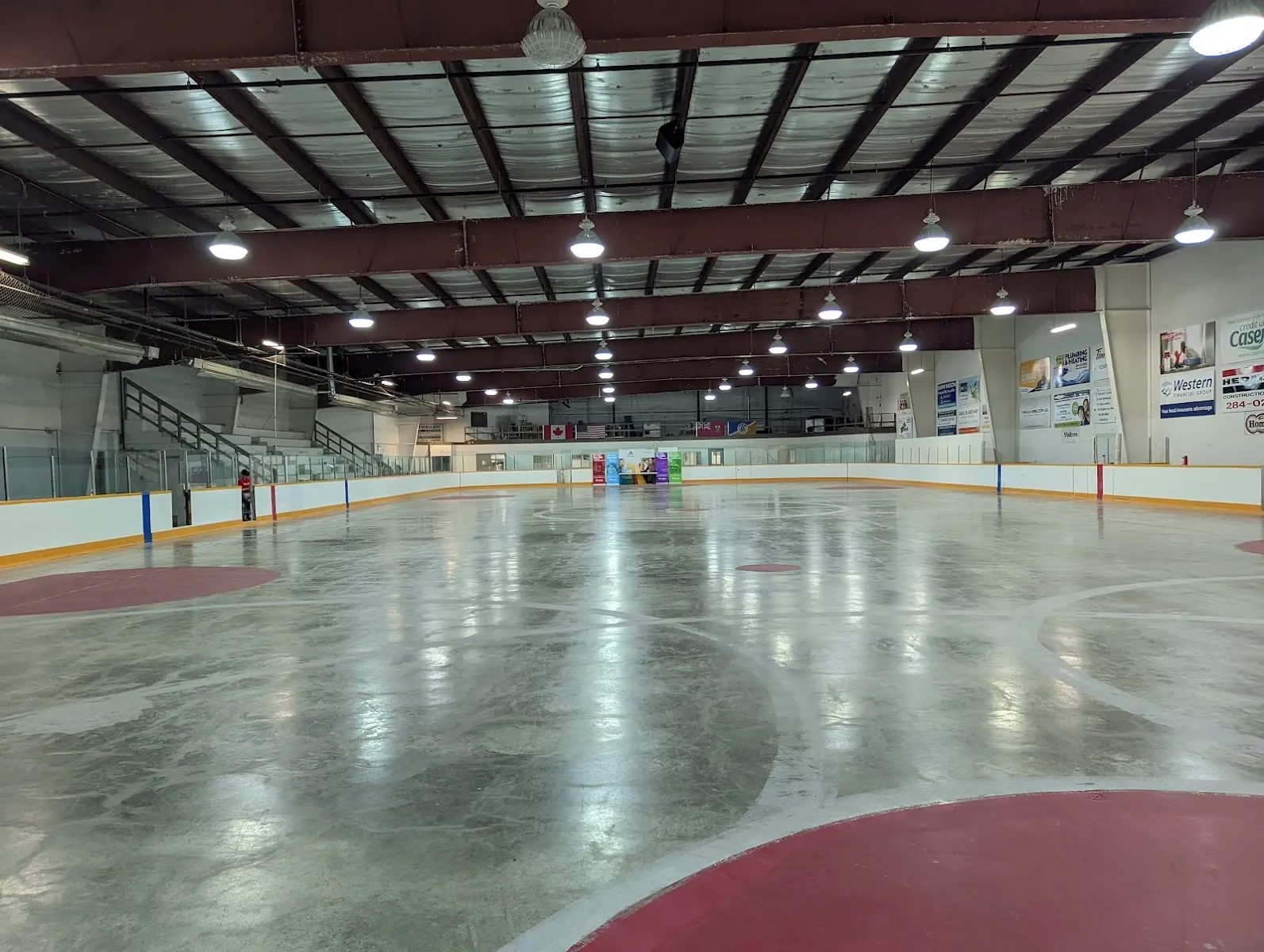 Una pista vacía con círculos de saque rojos, vallas blancas con detalles azules y amarillos y armaduras oscuras en el techo sobre hileras de luces. — Transcona East End Arena
