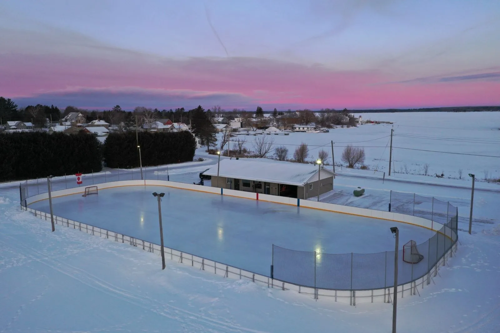 Aerial view of the Tranter Memorial Park outdoor rink at dusk, a boarded sheet of ice with a small shelter beside it and a frozen bay and shoreline houses behind. — Tranter Memorial Park Outdoor Rink (Richards Landing)