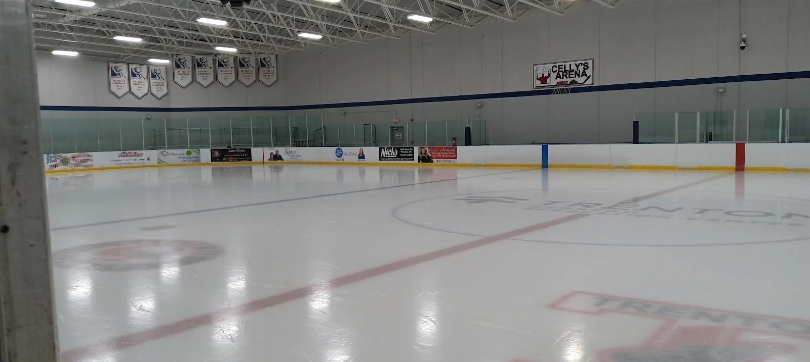 The empty sheet seen low from the near corner, white ice with red and blue lines and a lit wall of boards and glass along the far side. — Trenton Kennedy Recreation Center