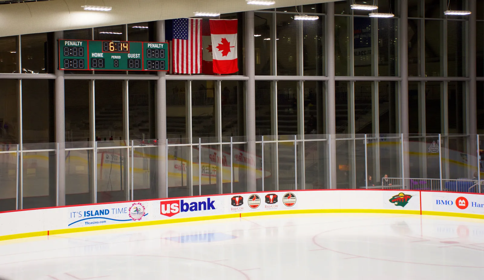 The empty sheet in front of the tall glass wall, with the scoreboard, U.S. and Canadian flags and sponsor boards along the dasher. — TRIA Rink
