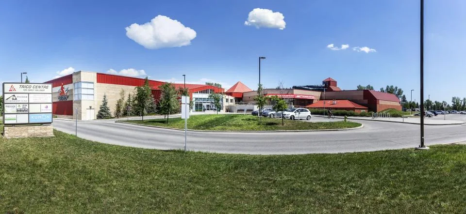Panorama of the red-roofed brick centre behind a landscaped roundabout, with the Trico Centre sign at the left and the parking lot beyond. — Trico Centre for Family Wellness