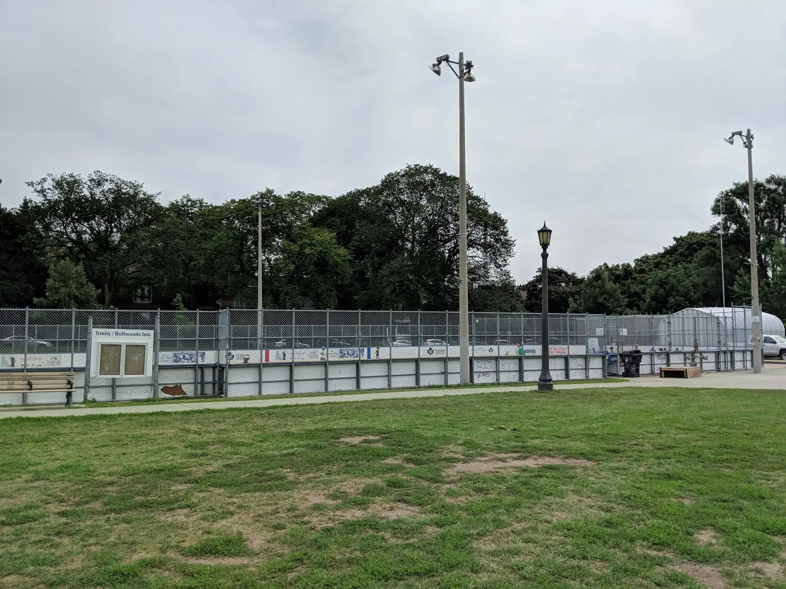 The outdoor rink's boards, fencing and floodlights at Trinity Bellwoods Park in summer, seen across the grass, with no people in view — Trinity Bellwoods Park Outdoor Ice Rink