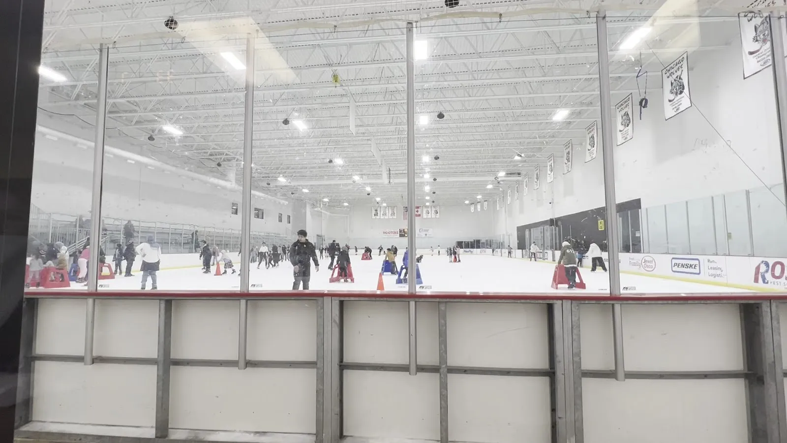 Una pista de hielo vista a través del cristal sobre las bandas, con patinadores en el hielo, cerchas de acero en el techo y banderines en la pared del fondo. — Triphahn Center Ice Arena