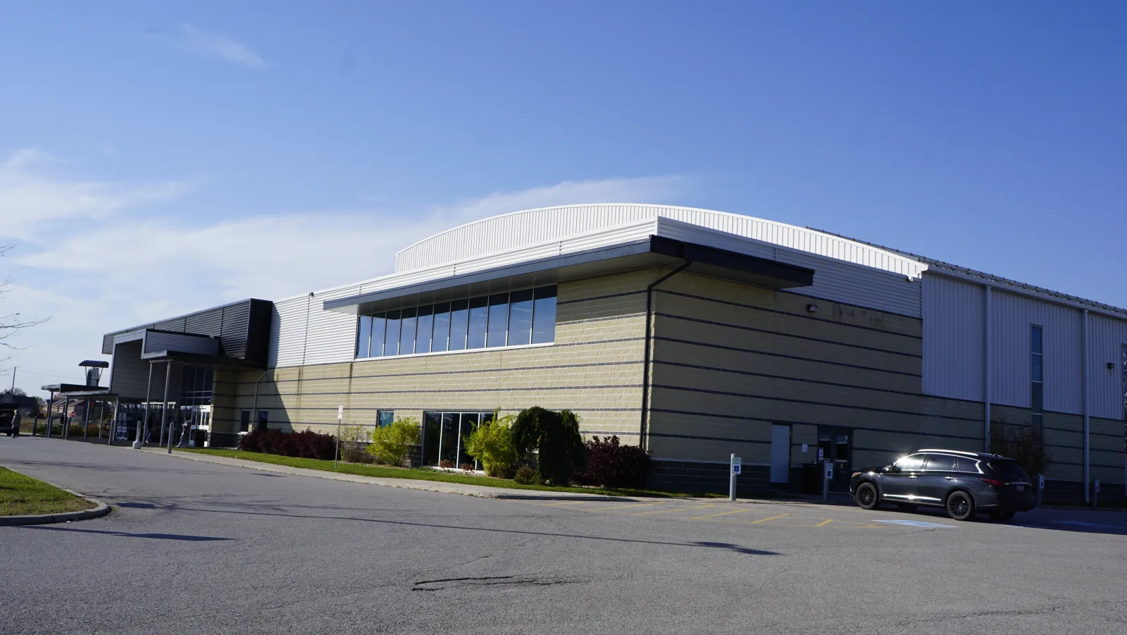 El largo costado de bloques y metal del Trisan Centre bajo un techo curvo, con una franja de ventanas en lo alto y la marquesina de la entrada a la izquierda. — Trisan Centre