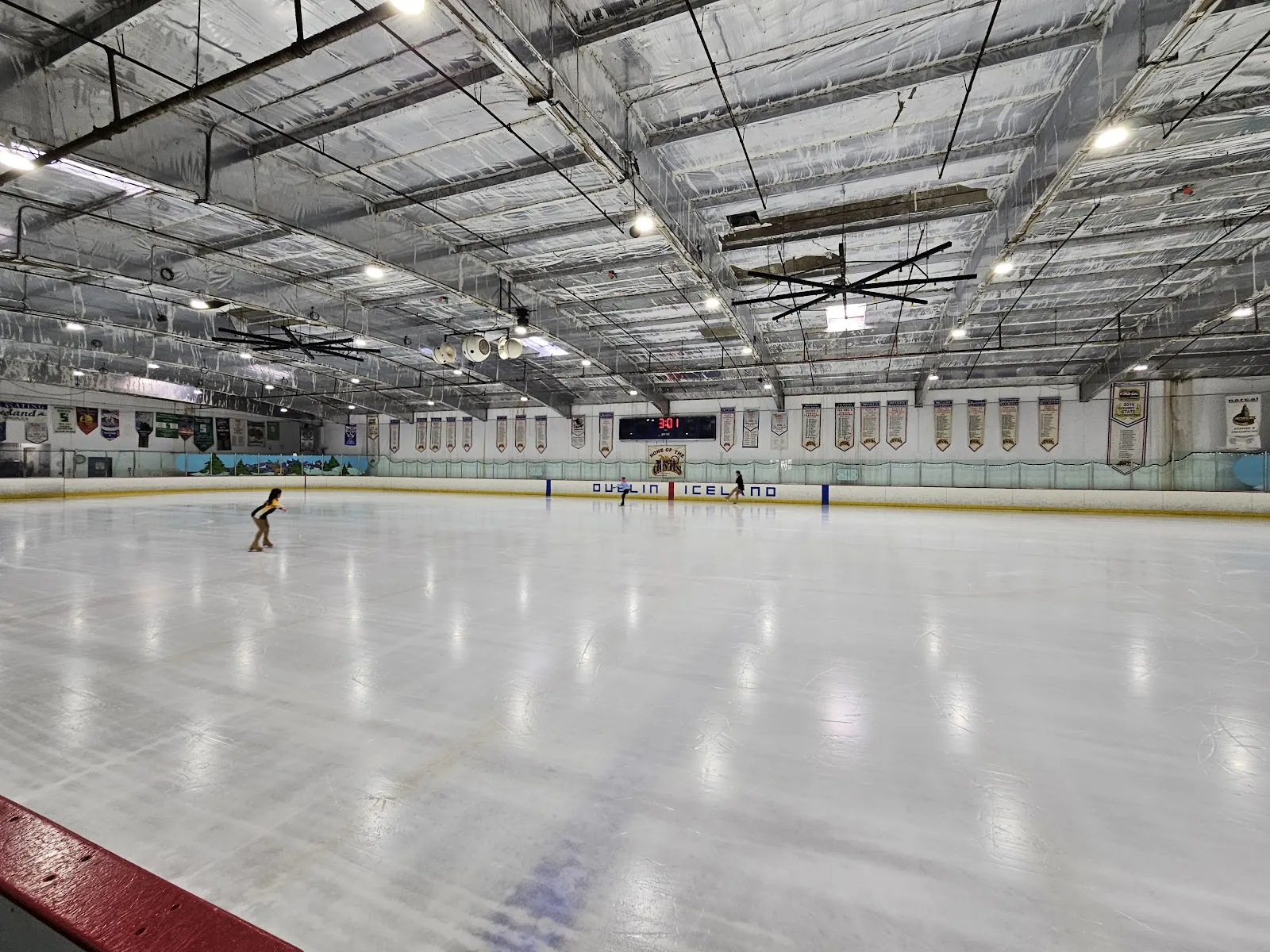 La amplia pista de Tri Valley Ice, recién pulida, con un patinador a lo lejos, bajo un techo con armaduras de acero e hileras de luces. — TriValley Ice Dublin