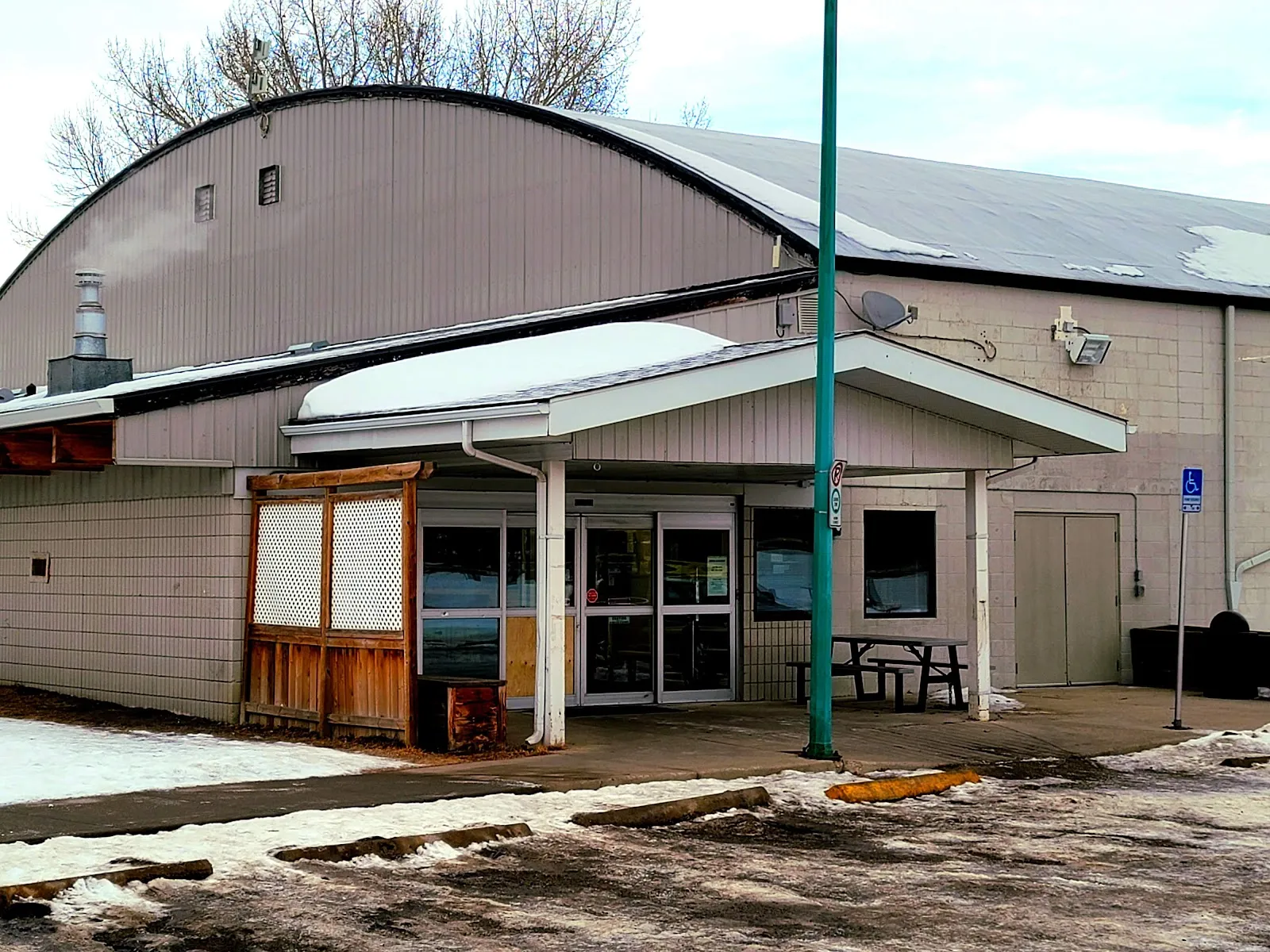 The arena's barrel roof above a covered entrance with glass doors, a wood-slatted patio screen and an accessible-parking sign, with snow on the ground. — Triwood Arena