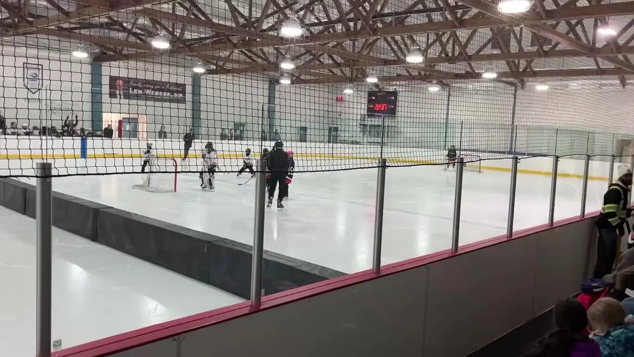 The sheet seen along the boards from the stands during a youth practice, with the scoreboard and banners on the far wall under wooden roof trusses. — Triwood Arena
