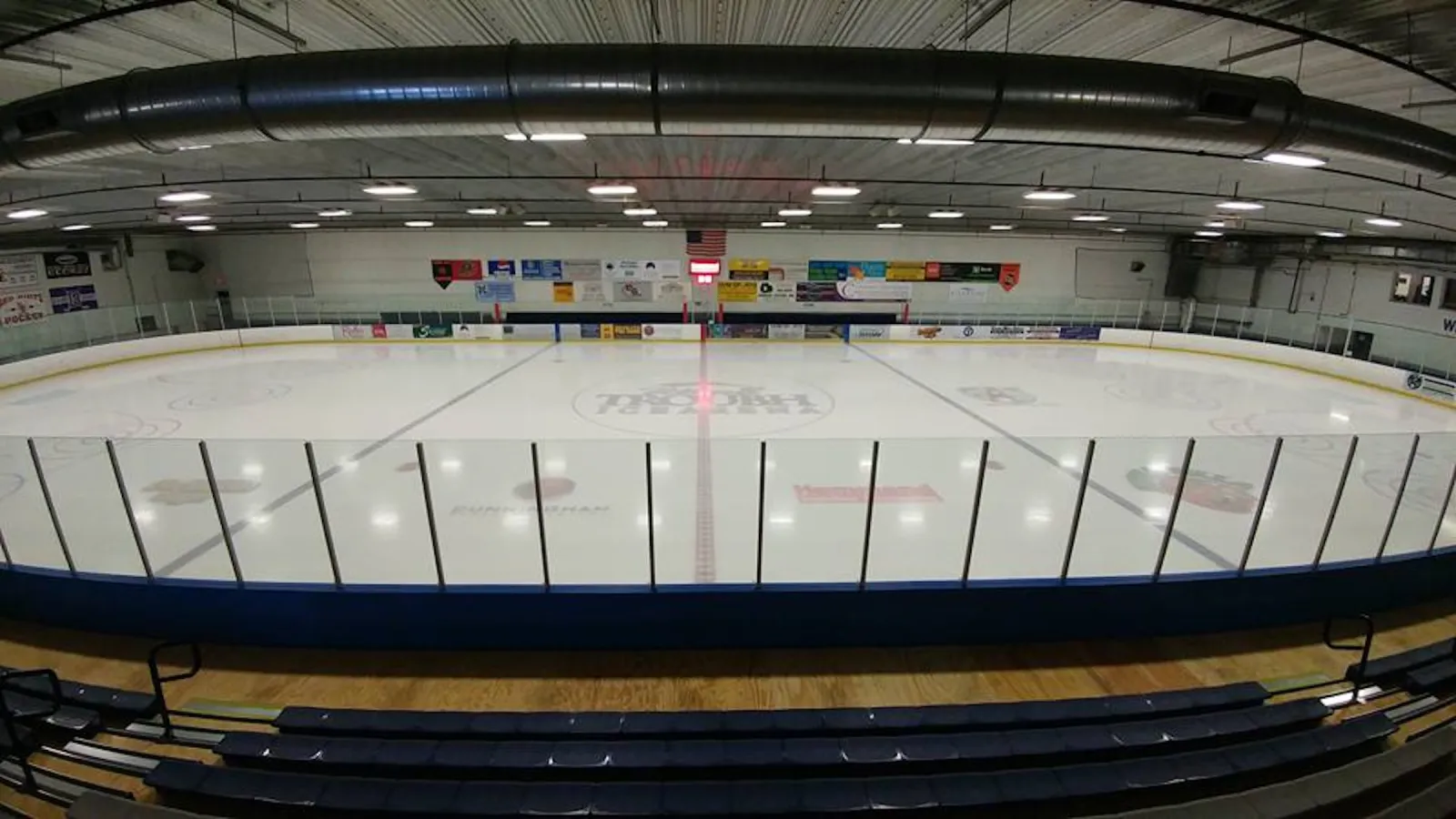 Empty NHL-size ice sheet viewed from the stands, with the Troubh Ice Arena center-ice logo, dasherboard ads and an American flag on the far wall. — William B. Troubh Ice Arena