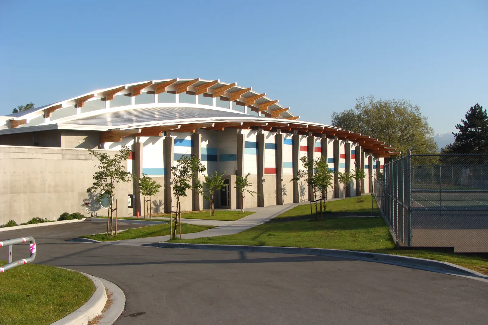 Entrada principal del edificio del Trout Lake Community Centre / Trout Lake Rink, con su marquesina curva de madera y acero, columnas de concreto en la entrada y las canchas de tenis contiguas, fotografiada desde el estacionamiento. — Trout Lake Rink