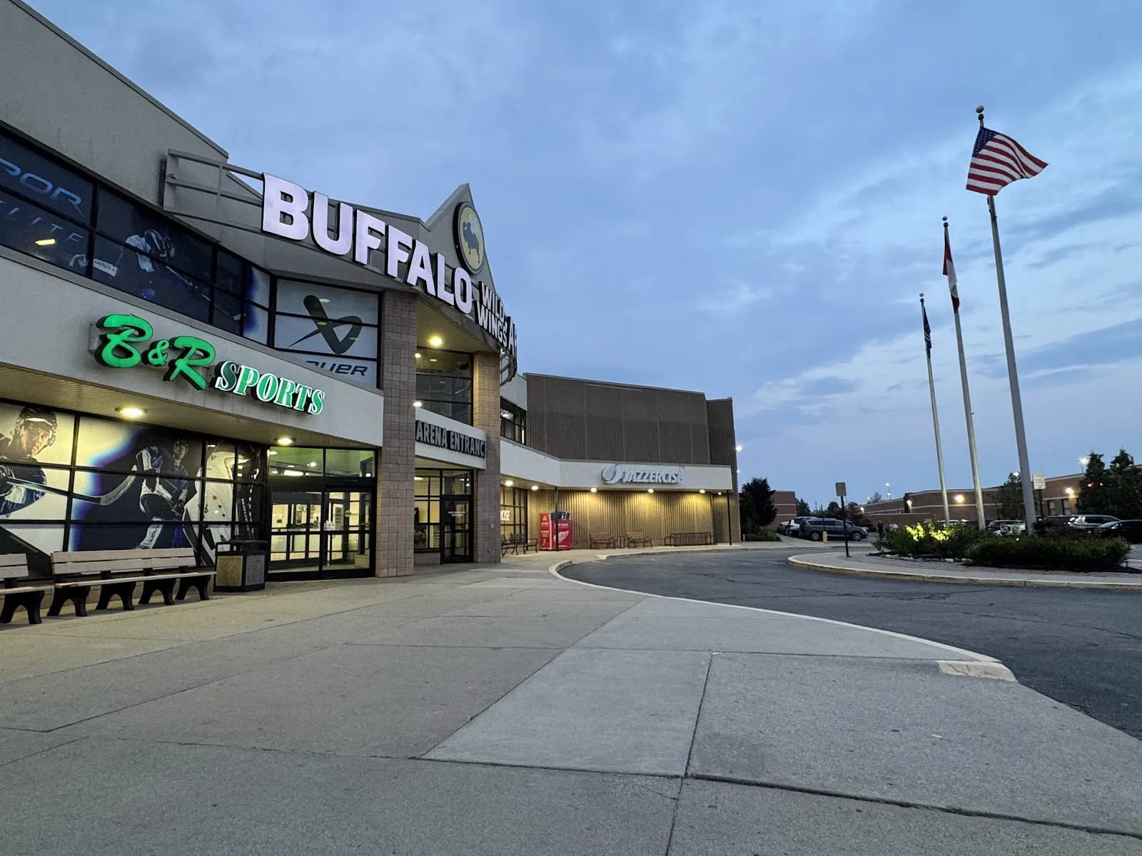 La entrada de Buffalo Wild Wings Arena al atardecer, con su nombre en letras grandes sobre las puertas de cristal y astas de bandera junto al estacionamiento. — Buffalo Wild Wings Arena