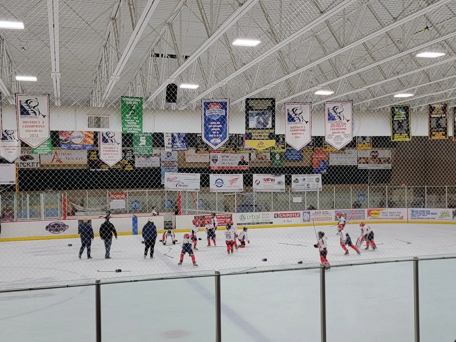 Una práctica bajo hileras de banderines del club, con jugadores y entrenadores repartidos por el hielo detrás de un barandal, y un largo techo claro arriba. — Buffalo Wild Wings Arena