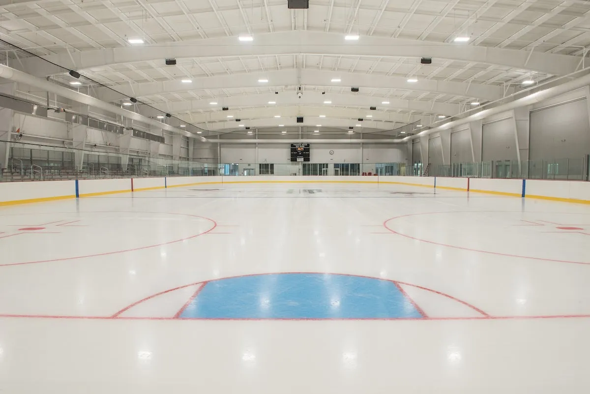 Pista vacía de tamaño NHL en Tucker Road Ice Rink, vista desde el nivel del hielo hacia las vallas del fondo, el marcador y las gradas para espectadores bajo un techo abovedado de armaduras blancas. — Tucker Road Ice Rink