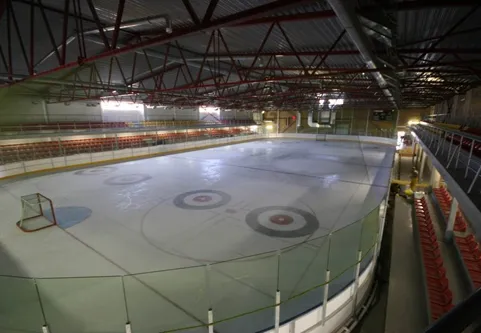 The empty ice sheet of the Tukuma ledus halle seen from the stands, with a hockey goal, curling circles painted on the ice and red seating along the far side under a steel roof. — Tukuma ledus halle