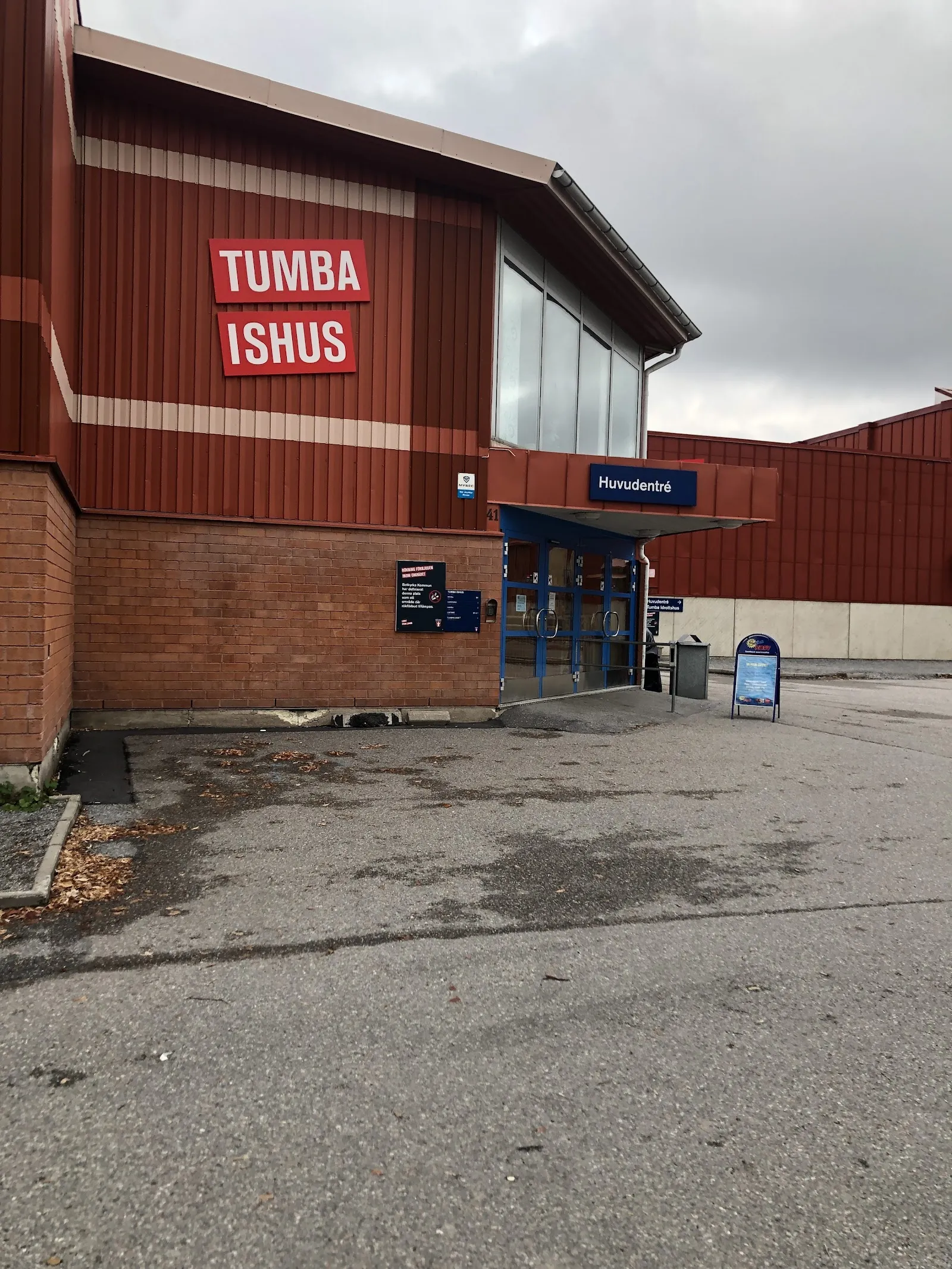 Tumba Ishus röda byggnad med namnskylt och den blå huvudentrén över en tom parkeringsplats – Tumba Ishus (hall 2)
