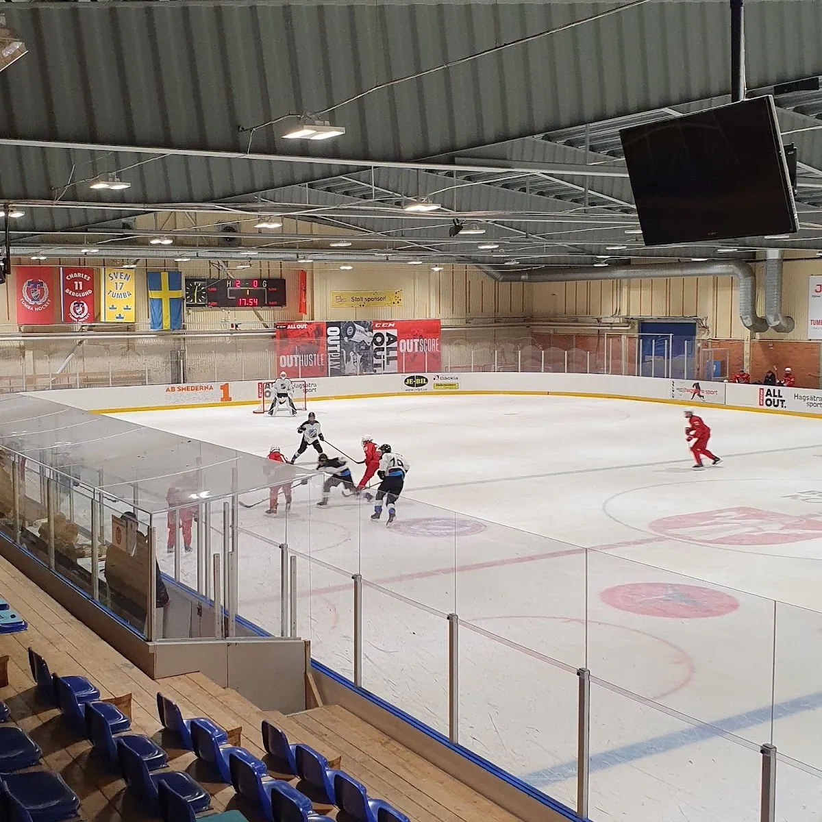 En match på isen i Tumba Ishus, sedd från läktaren över isytan med resultattavlan hängande ovanför och spelarna nära sargen. – Tumba Ishus (hall 2)