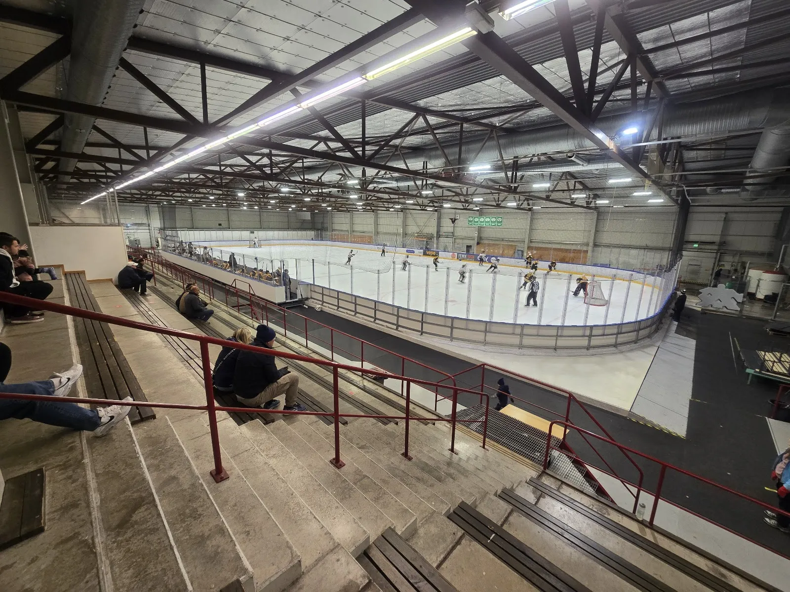 Isytan i Tuusulan jäähalli sedd från betongläktaren, inne i hallen med stålfackverk, sarg och nät, några små spelare långt borta på isen och en del åskådare på läktaren – Tuusulan jäähalli (Hyrylä)