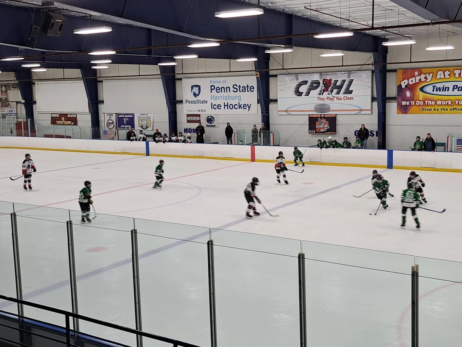 Un partido visto a través del vidrio, con jugadores repartidos por una pista amplia bajo un techo claro con banderines de Penn State y de la liga en la pared del fondo. — Twin Ponds Arena