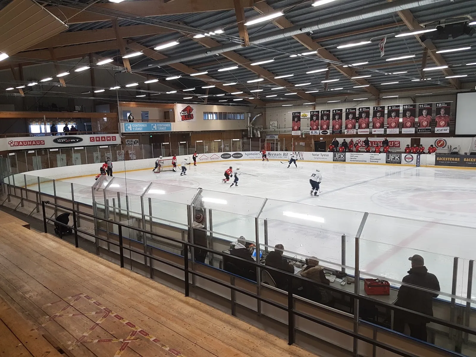 Players on the ice at Tyresö ishall seen from the wooden stand, with the timber roof and sponsor boards behind. — Tyresö ishall