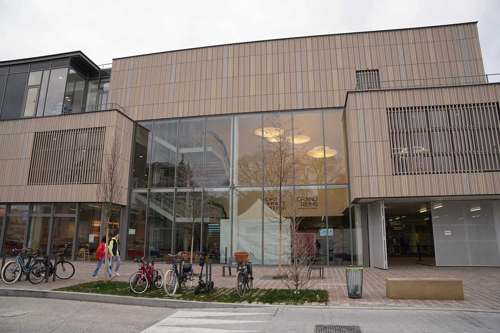 La façade vitrée de l’entrée de l’UCPA Sport Station Grand Reims, rue Jules César, avec les enseignes Sport Station et Grand Reims sur le verre et quelques personnes et vélos au pied du bâtiment. — UCPA Sport Station Grand Reims