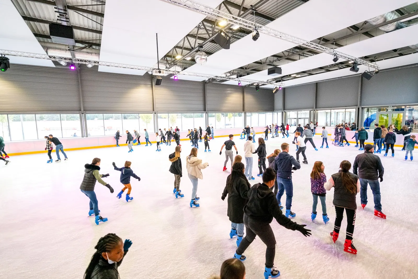 Patinage public dans la grande halle à toit de verre : une foule de patineurs de tous âges répartie sur la glace, sous le toit métallique éclairé. — UCPA Sport Station Grand Reims