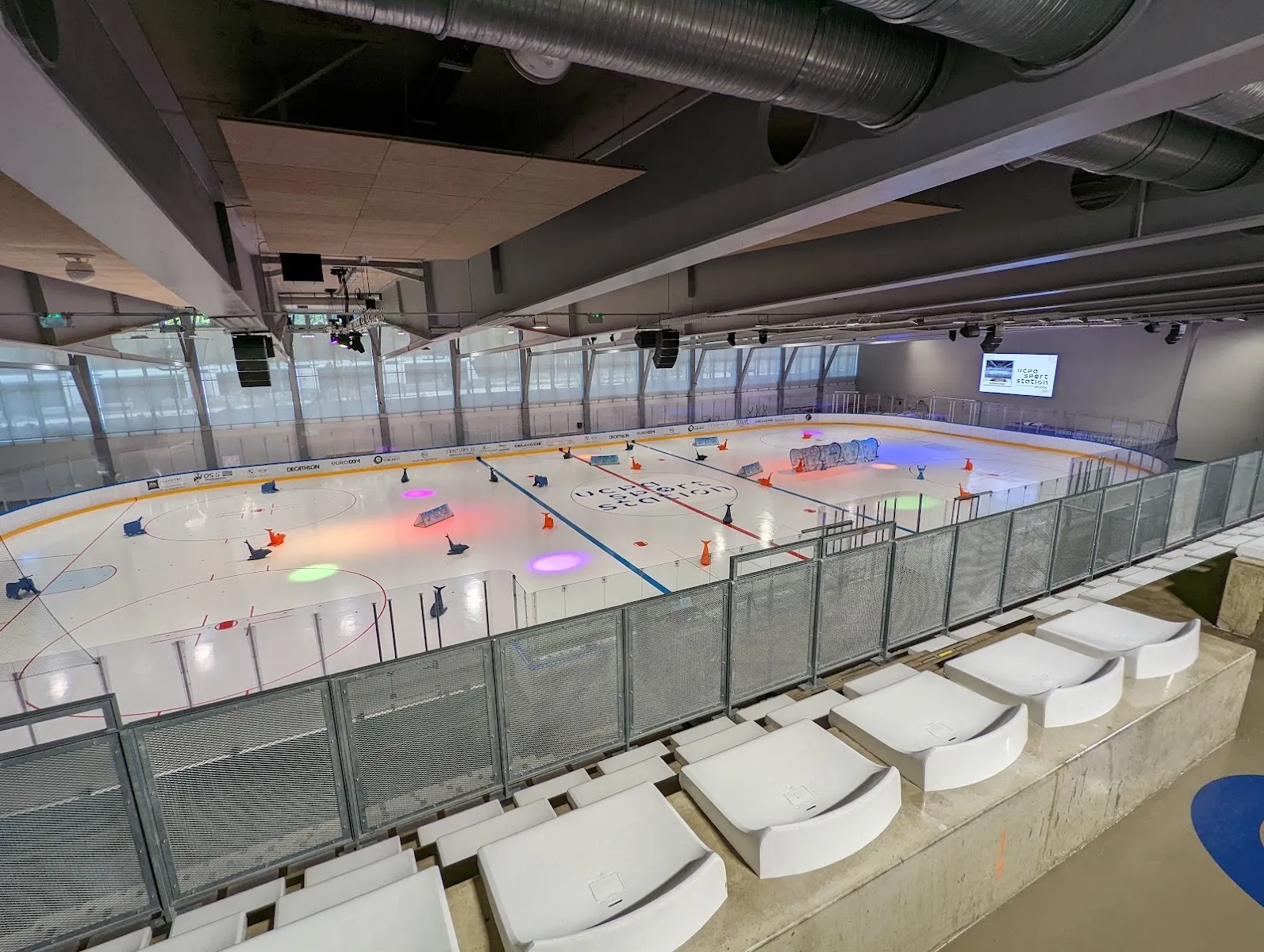 Raised view over the Meudon ice from the stands, with the sheet marked out in coloured training zones and cones, the steel roof structure and a row of white stands in the foreground; no skaters in the foreground. — UCPA Sport Station Meudon