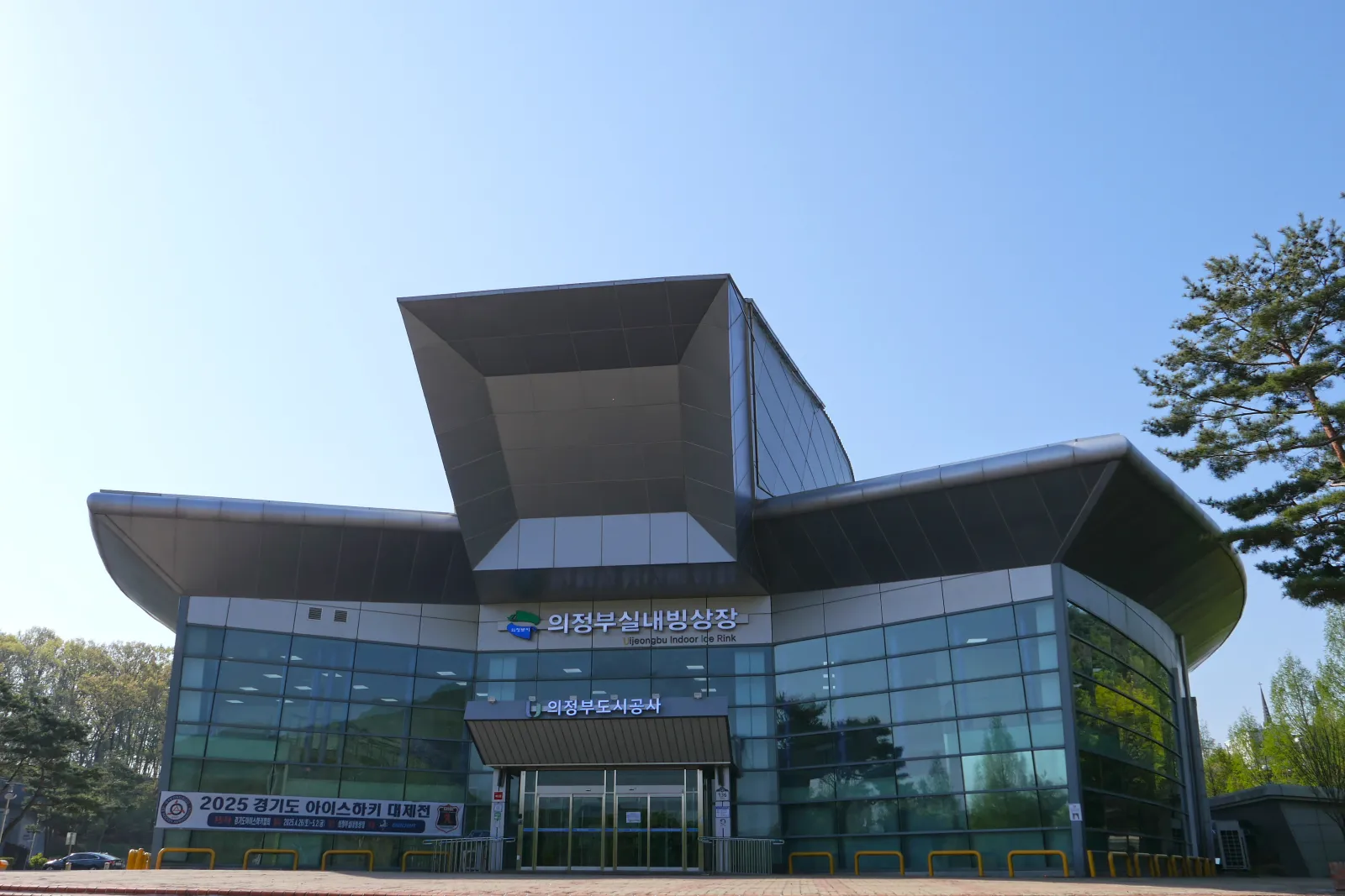 The glass-fronted entrance of Uijeongbu Indoor Ice Rink under its angular steel canopy, with the hall's name over the doors, seen from the forecourt. — Uijeongbu Indoor Ice Rink